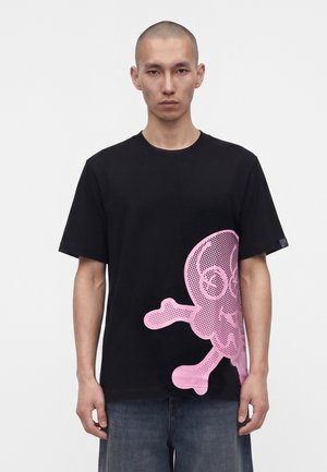 Ung mand iført en sort t-shirt med en stor pink dødningehoved- og korsknoglegrafik på nederste højre side, kombineret med blå jeans.