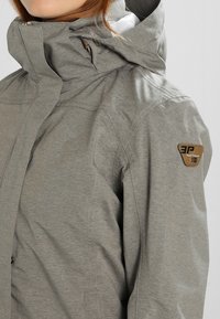 Veste gris clair avec capuche, en tissu texturé, fermeture éclair, et patch logo triangulaire sur la manche gauche.