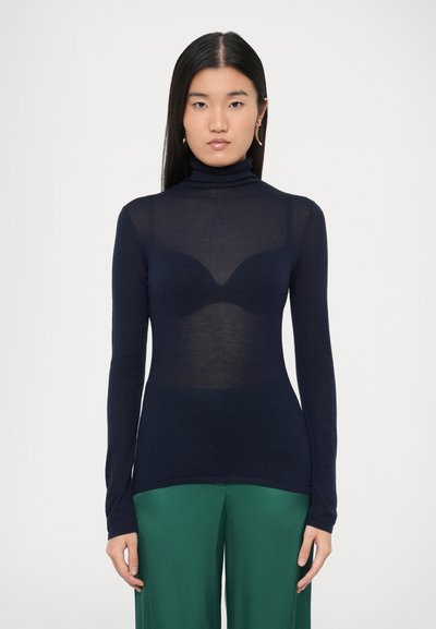 Haut à manches longues col roulé bleu marine en tissu transparent, avec un design subtil de contour de buste. Associé à un pantalon vert à jambes larges.