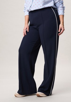 Persoon die marineblauwe wijde broek met witte zijstreep, lichtblauw overhemd met rood hartjespatroon en beige sneakers draagt, staand met ontspannen handen.