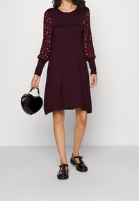 Robe tricotée bordeaux avec des manches bouffantes texturées ornées de points dorés. Assortie à un sac à main noir en forme de cœur et des chaussures Mary Jane à patin noir.