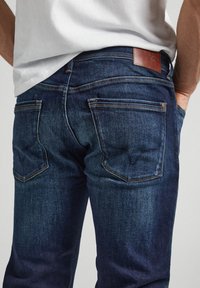 Jeans de mezclilla azul oscuro con una textura suave, cinco bolsillos y una etiqueta de cuero en la parte trasera de la cintura. Presenta detalles de costura en contraste.