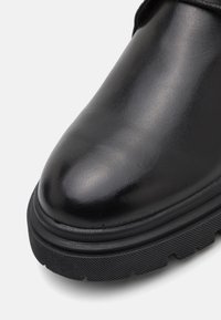 Bota de cuero negro con un acabado suave y brillante, punta redondeada y una suela de goma texturizada y gruesa. Presenta costuras sutiles y sin hardware visible.