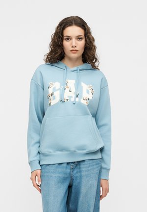 Tânără cu păr creț purtând un hanorac GAP bleu cu imprimeu floral și blugi albaștri, pe un fundal alb simplu.