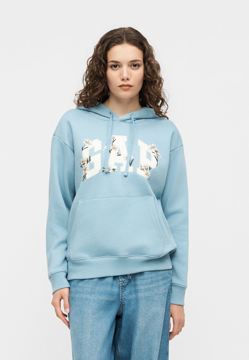 Νεαρή γυναίκα με σγουρά μαλλιά, φορώντας ένα ανοιχτό γαλάζιο φούτερ GAP με λουλουδάτο σχέδιο και μπλε τζιν, σε λευκό φόντο.