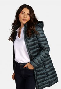 Manteau long rembourré en bleu sarcelle avec une finition brillante, doté d'une capuche, d'une fermeture éclair à l'avant et d'une silhouette ajustée. Porté par-dessus un polo blanc.