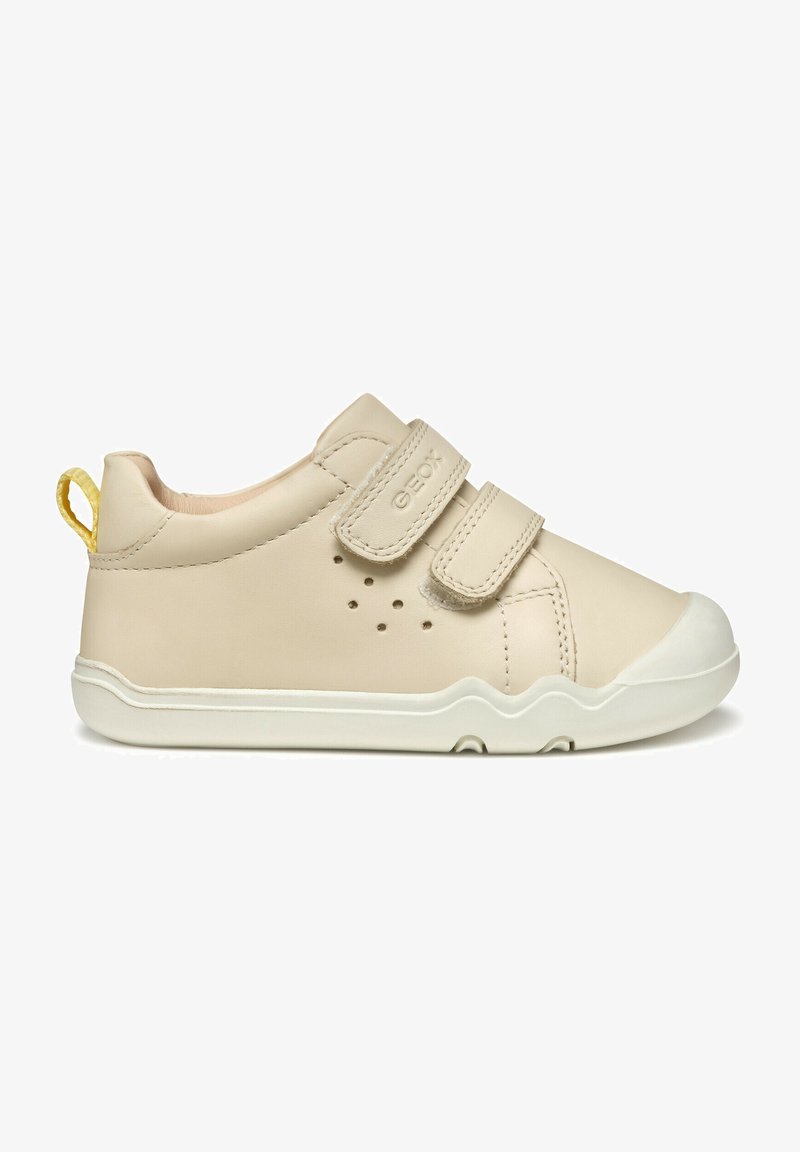 Basket beige en cuir avec deux bandes Velcro, bout arrondi et semelle en caoutchouc blanche. Présente de petits trous de ventilation sur le côté et un tirant au talon jaune.
