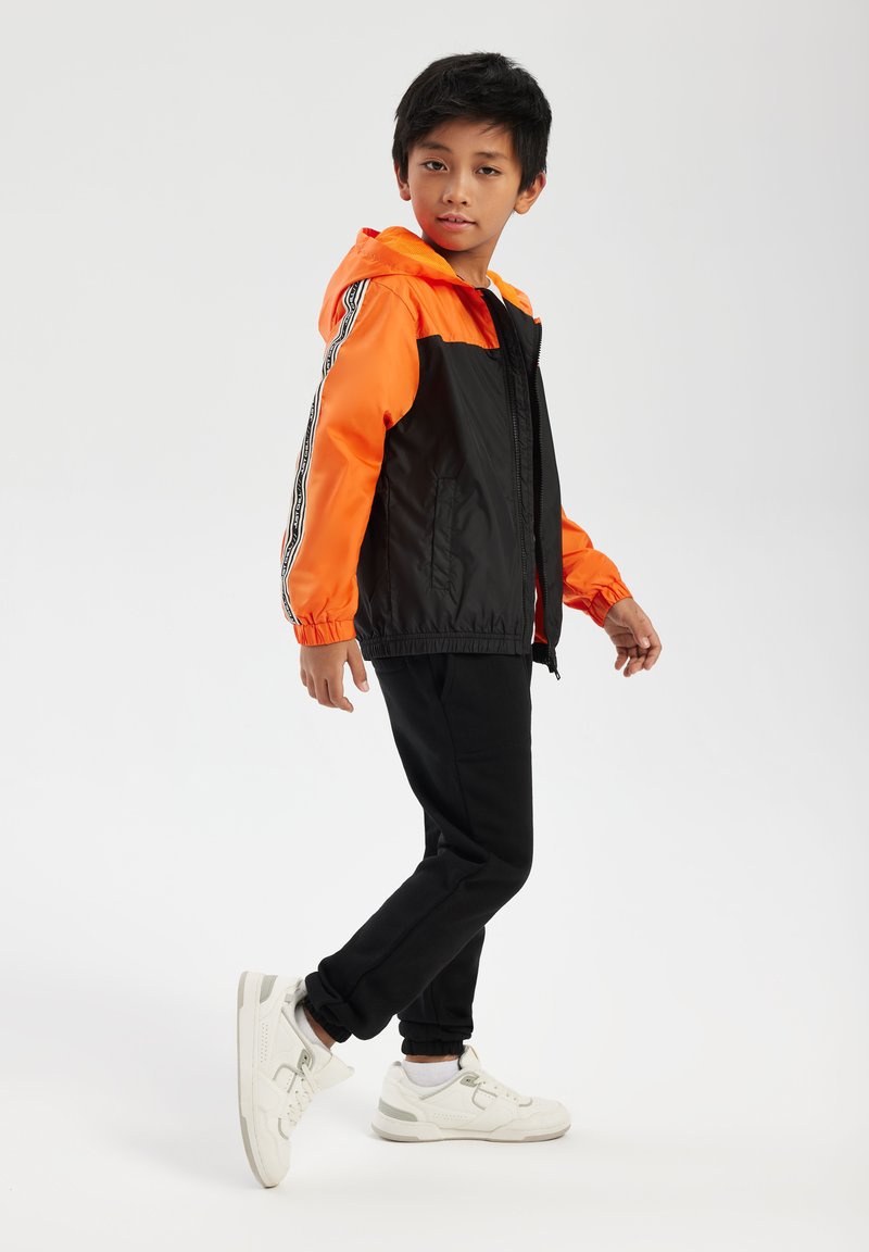 Zwarte en oranje windbreaker met capuchon, met contrasterende mouwen, elastische manchetten en een rits aan de voorkant. Gekleed met zwarte trainingsbroek en witte sneakers.