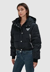 Czarna kurtka puchowa o skróconym kroju, z odpinanym kapturem i zatrzaskami. Posiada haftowane logo oraz kontrastowy napis po boku.