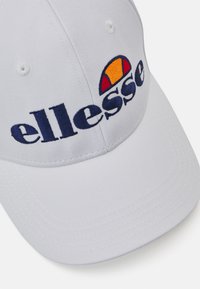 Biała czapka baseballowa z haftowanym logiem "ellesse" w kolorze granatowym oraz kolorową grafiką pół-słońca. Gładka tekstura i dopasowany krój.