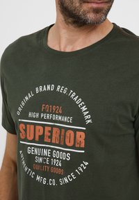 Mørkegrønn bomull t-skjorte med rund hals, med hvit og oransje grafisk trykk som sier "SUPERIOR" og andre tekstelementer i et sirkulært design.