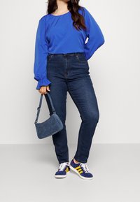 Jeans de mezclilla de cintura alta, combinados con una blusa larga de color azul vibrante con mangas abullonadas. Los accesorios incluyen un pequeño bolso azul y zapatillas deportivas.