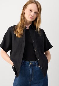 Svart kortärmad skjorta med knapplist, fickdetalj och linneaktig textur, kombinerad med högmidjade blå denimjeans.