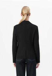 Blondinööriga naine, kellel on seljas sobiv must bleiser ja tumesinised teksad, seisab näoga ära lihtsa valge tausta vastu.