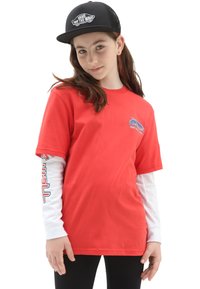 T-shirt in cotone rosso con maniche corte, indossato sopra una camicia bianca a maniche lunghe. Presenta un logo grafico sul petto. Cappellino nero con logo.