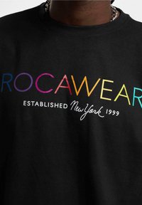 Juodas medvilninis marškinėliai su "ROC AWEAR" daugiavardžiais rašmenimis, o po juo baltu kreivuoju šriftu užrašas "ESTABLISHED NewYork 1999".