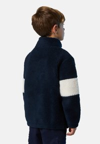 Niño pequeño de pie de lado, vistiendo una chaqueta de forro polar azul marino con una franja horizontal blanca en las mangas, frente a un fondo liso.