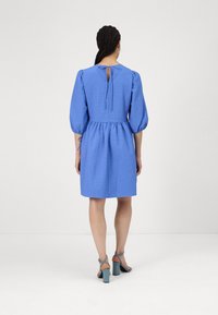 Kék, rövid ruha puffos ujjal, texturált anyagból, szűkített derékkal és kulcslyuk formájú hátkivágással. Kék, vastag sarkú szandált visel.