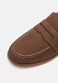 Bruna mockaskor med en sydd design, med ett dekorativt band över ovandelen och en slät, rund tå.