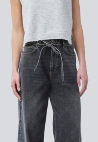 Frau in hoch taillierten schwarzen Jeans mit einem gebundenen Stoffgürtel und einem grauen ärmellosen, kurz geschnittenen Pullover.