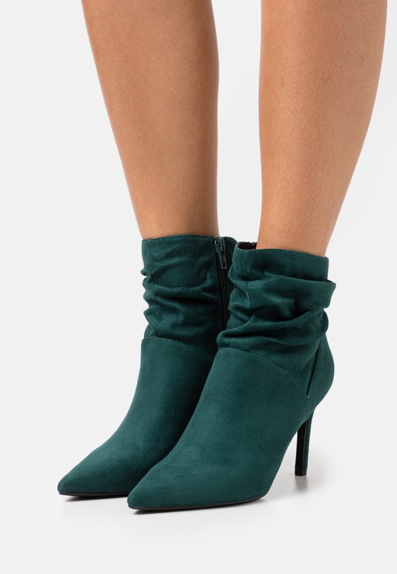 Femme portant des bottines en daim vert foncé à bout pointu, tige souple et talons aiguilles hauts sur fond blanc.