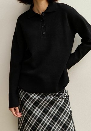 Person trägt einen schwarzen Polo-Pullover mit langen Ärmeln und einen schwarz-weiß karierten Rock, steht mit einer Hand in der Hüfte.