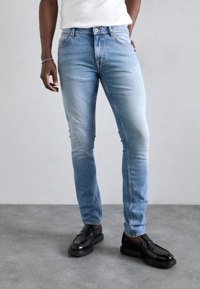Világoskék farmernadrág, slim-fit szabásún, finom kopással és klasszikus öt zsebes stílussal. Fekete bőrcipővel viselve.