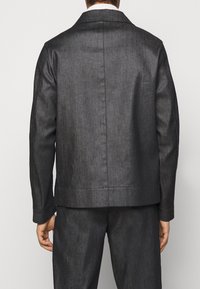 Chaqueta gris oscuro hecha de tela texturizada, con cuello muesca, mangas largas y dobladillo recto. Detalles de costura visibles en los hombros.