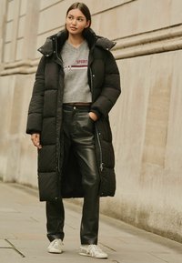 Cămașă puffer neagră cu glugă, pulover tricotat gri cu textul "SPORTS", pantaloni din piele negri și adidași albi cu accente deschise.