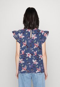Blumenbluse in Marineblau mit Rüschensäumen, die pinke und lila Blumenmuster zeigt, sowie einem kleinen Schlüsselaugen-Detail auf der Rückseite.