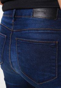 Jeans de mezclilla azul oscuro con una textura suave, que presentan costuras en contraste, dos bolsillos traseros y una etiqueta de cuero negro en la cinturilla.