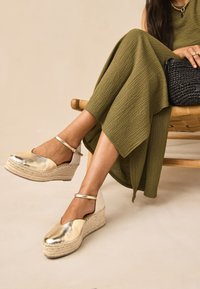 Guld kilklackssandaler med texturerad yta, utrustade med en ankelrem och en sula av flätad jute. Matchas med en olivgrön klänning.