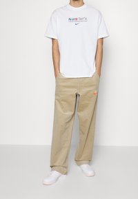 Vit kortärmad t-shirt med färgglad text och Nike-logotyp, tillsammans med beige kordbyxor med en orange Nike-emblemet, och vita sneakers.