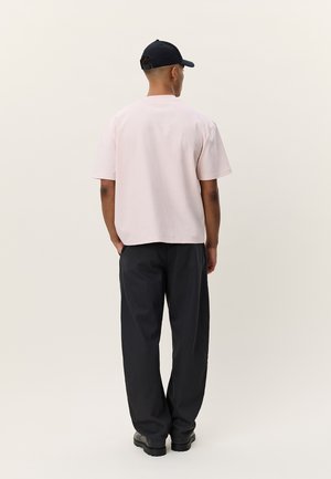 Ροζ κοντομάνικο μπλουζάκι με άνετη γραμμή, συνδυασμένο με φαρδιά μαύρη παντελόνια και μπλε σκούφο, από πίσω, σε ουδέτερο φόντο.