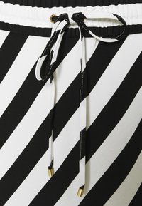 Musta-valkoraidallinen kangas, jossa kiristysnyöri vyötäröllä ja kullanväriset nyörinpäät sekä musta kontrastitikkaukset. Sileä pinta ja jämäkkä muoto.