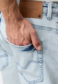 Svijetloplave traperice s vidljivim stražnjim džepom i kožnom oznakom branda. Imaju detalje šivanja i izlizanu teksturu. Ruka se odmara u džepu.