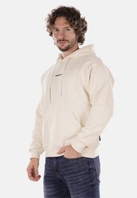 Felpa con cappuccio di colore crema, dotata di una tasca frontale e testo del logo nero. Presenta maniche lunghe e una vestibilità comoda, abbinata a jeans di jeans scuri.