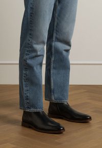 Schwarze Leder-Chelsea-Stiefel mit spitz zulaufender Zehe, getragen mit hellblauen Jeans, auf einem Holzboden. Sichtbarer seitlicher Elastikeinsatz und flacher Absatz.