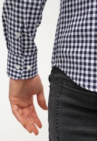 Camicia a quadri blu e bianchi con polsini con bottoni, realizzata in tessuto di cotone liscio. Mano appoggiata accanto a un pantalone di denim di colore scuro.