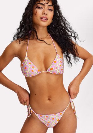 Femme aux longs cheveux bouclés portant un bikini triangle rose à motif floral, ajustant les liens du bas de bikini des deux côtés.