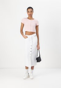 Lys rosa cropped t-skjorte med knappedetaljer, kombinert med en hvit denim skjørt med knapper foran og hvite sneakers. En svart håndveske gir kontrast.