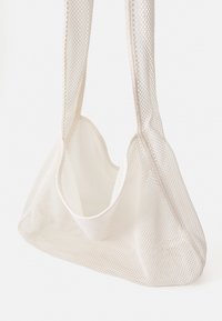 Sac en filet de couleur blanc cassé. Présente une forme douce et flexible avec de larges anses tissées et un compartiment ouvert spacieux pour un accès facile.