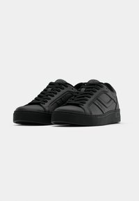 Paire de baskets noires basses avec semelles épaisses, lacets noirs et détails de panneaux cousus sur un fond clair et uni.