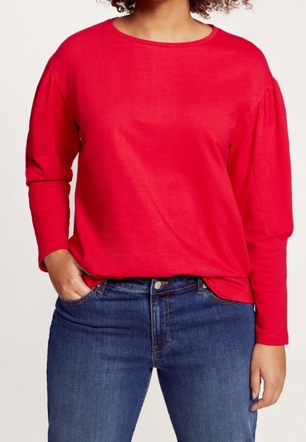 Punainen pitkähihainen paita, jossa on pyöreä kaula-aukko ja puffihihat, yhdistettynä sinisiin farkkuihin. Kangas näyttää pehmeältä ja sileältä.