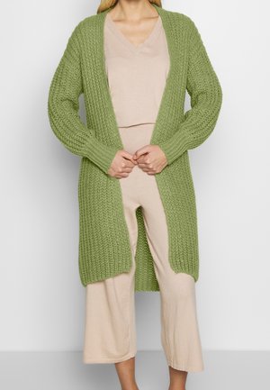 Kvinde iført lysebeige v-hals top og bukser med en lang, chunky strikket olivengrøn cardigan, stående mod en ensfarvet baggrund.