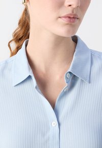 Donna che indossa orecchini a cerchio dorati e una maglietta azzurra a coste con il primo bottone slacciato, che mostra il collo e le labbra.