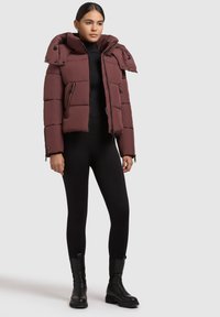 Burgundy prošívaná bunda s vysokým límcem a zipy, s horizontálním prošíváním a bočními kapsami. Nosená přes černý top a legíny.