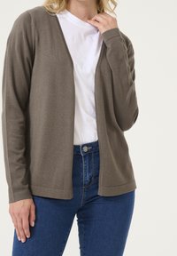Leichter brauner Cardigan mit V-Ausschnitt, langen Ärmeln und ohne Verschlüsse, kombiniert mit einem weißen Hemd und blauen Jeans.
