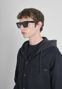 Crni preveliki dukserica s kapuljačom na vezanje, zatvaranjem na dugme i dva prednja džepa; nosi se preko crne majice; upotpunjena oštrim crnim naočalama.