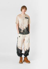 Camiseta de gran tamaño en un estampado desenfocado en beige y negro, con un bolsillo en el pecho, combinada con pantalones sueltos en crema y negro. Botas marrones con cordones.